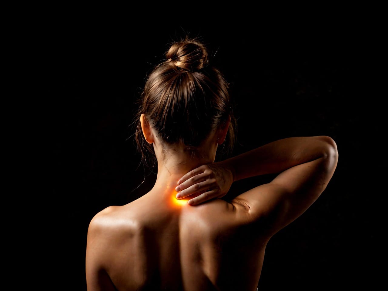 Woman with hair in a bun holding her upper back and neck in pain, highlighted with an orange glow to indicate muscle tension or injury, viewed from behind on a dark background.